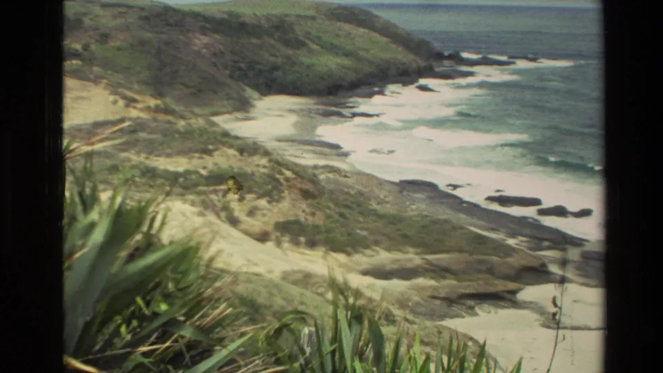 1981 New Zealand home-movie frame showing a rugged beach, dark rocks, surf, grassy foreground, and distant headland.