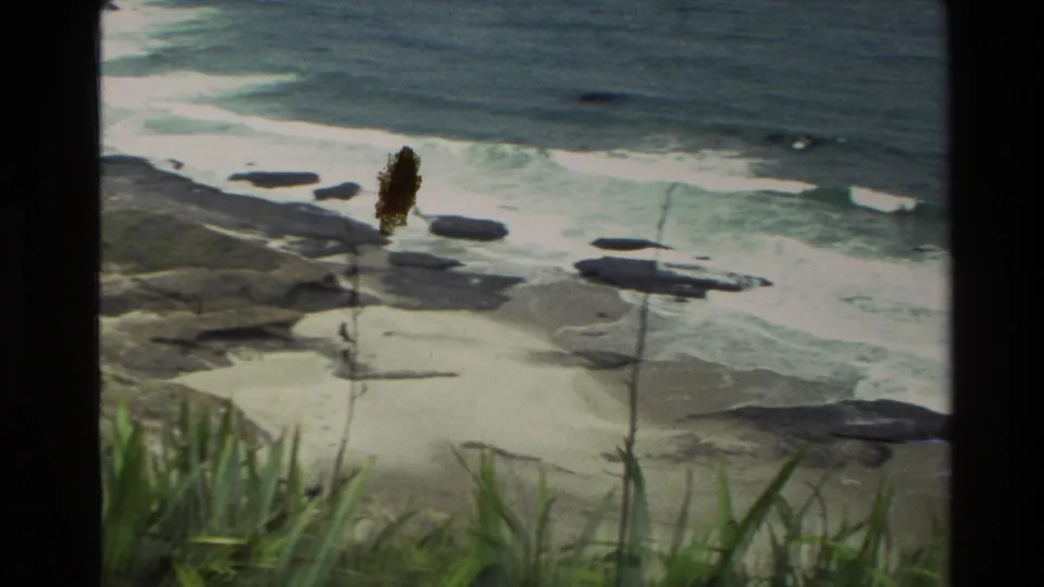 1981 New Zealand home-movie frame showing surf washing around dark rocks on a sandy coastal beach.