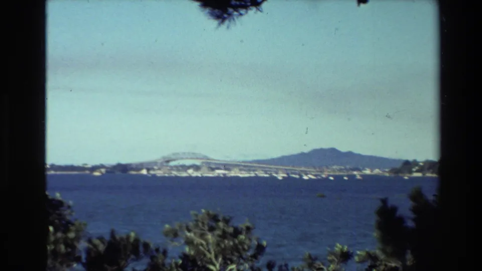 1981 New Zealand home-movie view across blue water toward a long bridge and distant hill, with trees framing the lower foreground.