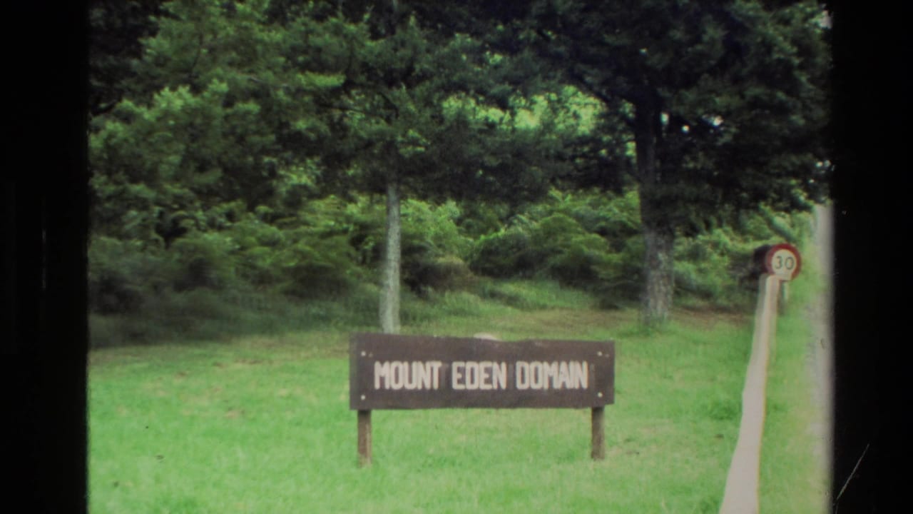 1981 New Zealand frame with a wooden Mount Eden Domain sign in a grassy park area, backed by trees and a small speed sign.
