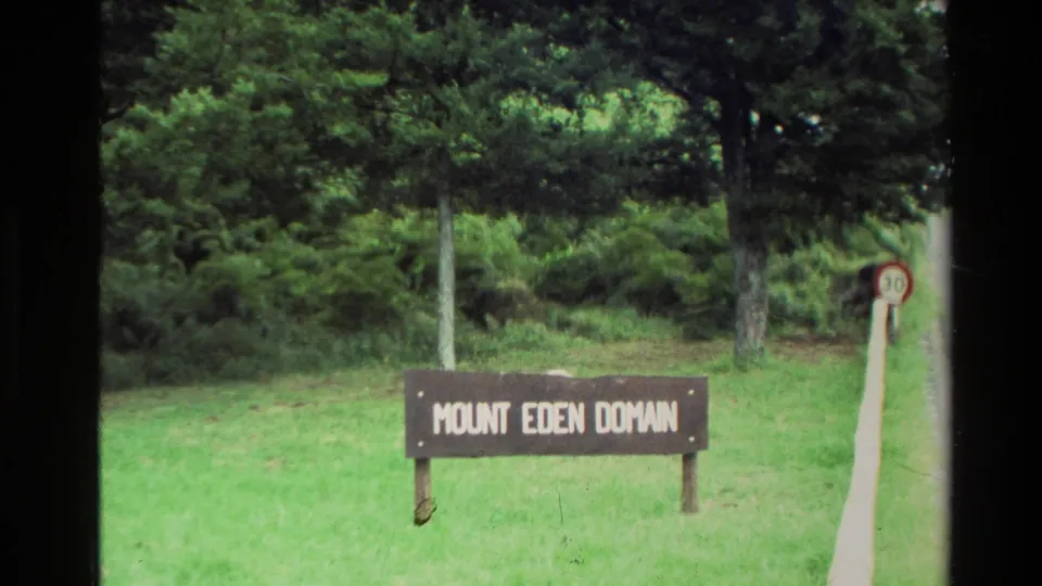 1981 New Zealand 8mm frame showing the Mount Eden Domain wooden sign, green lawn, trees, brush, and a small circular speed sign.