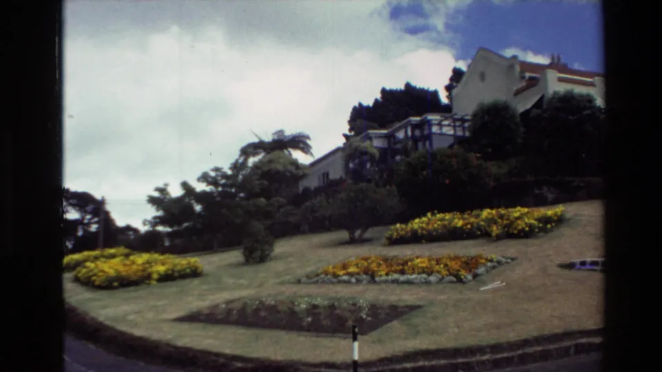 1981 New Zealand frame showing a large white building on a hillside above manicured lawns, yellow flower beds, trees, and clouded sky.