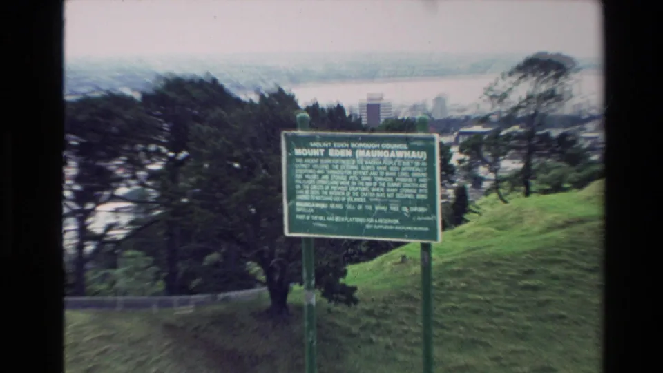 Mount Eden Maungawhau sign overlooking a grassy slope, trees, Auckland buildings, and harbor water.