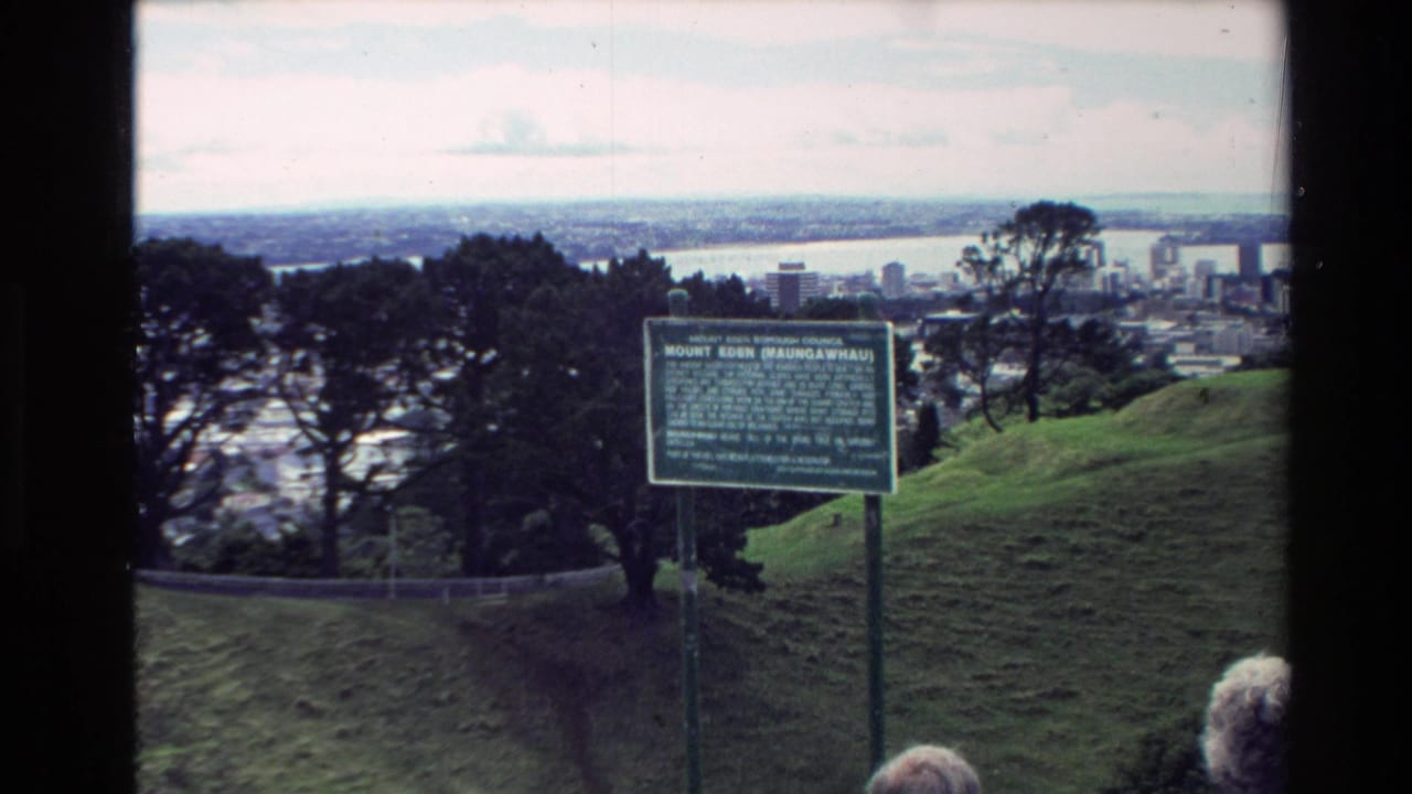 1981 New Zealand home-movie frame showing the Mount Eden Maungawhau sign, trees, Auckland skyline, and harbor from a grassy overlook.