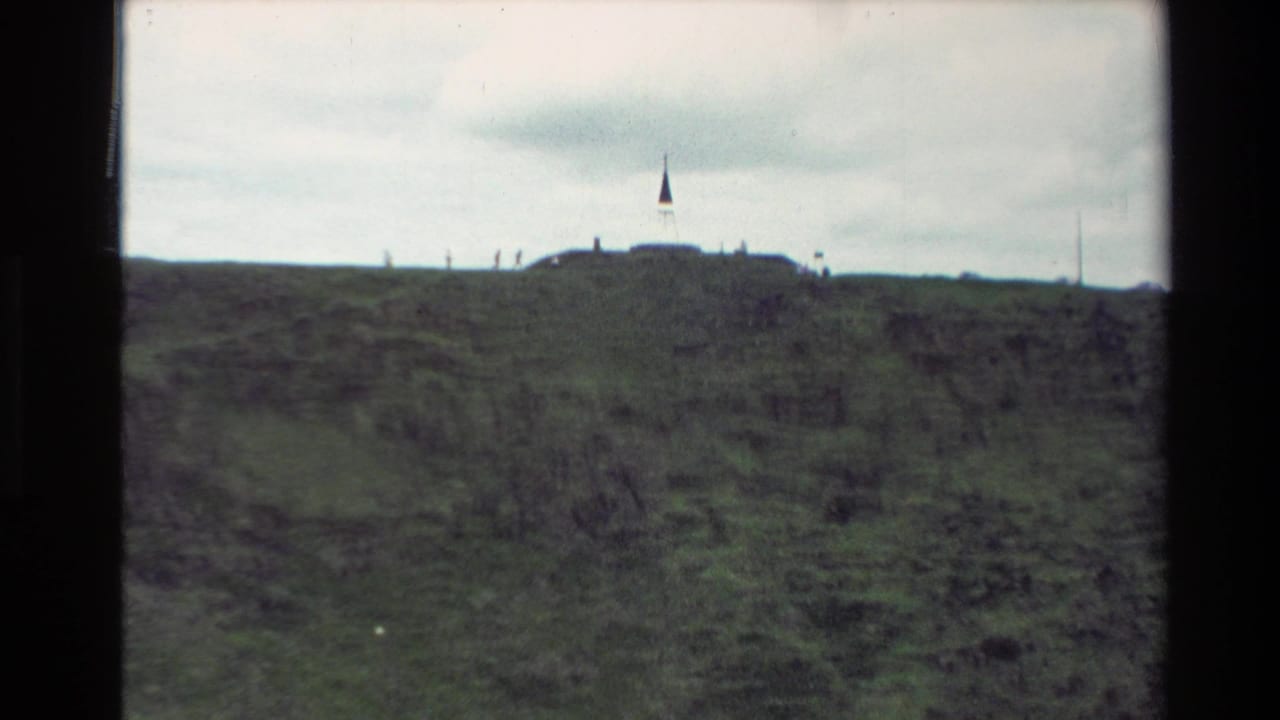 1981 New Zealand home-movie frame looking across the grassy Mount Eden hilltop toward small visitor figures and a flagpole on the ridge.