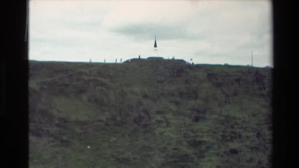 1981 New Zealand home-movie frame looking across the grassy Mount Eden hilltop toward small visitor figures and a flagpole on the ridge.