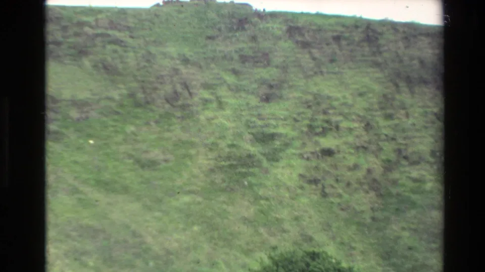 1981 New Zealand home-movie frame showing a soft, close view of the green Mount Eden slope with a pale sky edge and film gate border.
