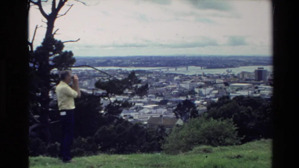 In 1981 New Zealand home-movie footage, a man in a yellow shirt uses binoculars from a grassy hill overlooking a coastal town and harbor.