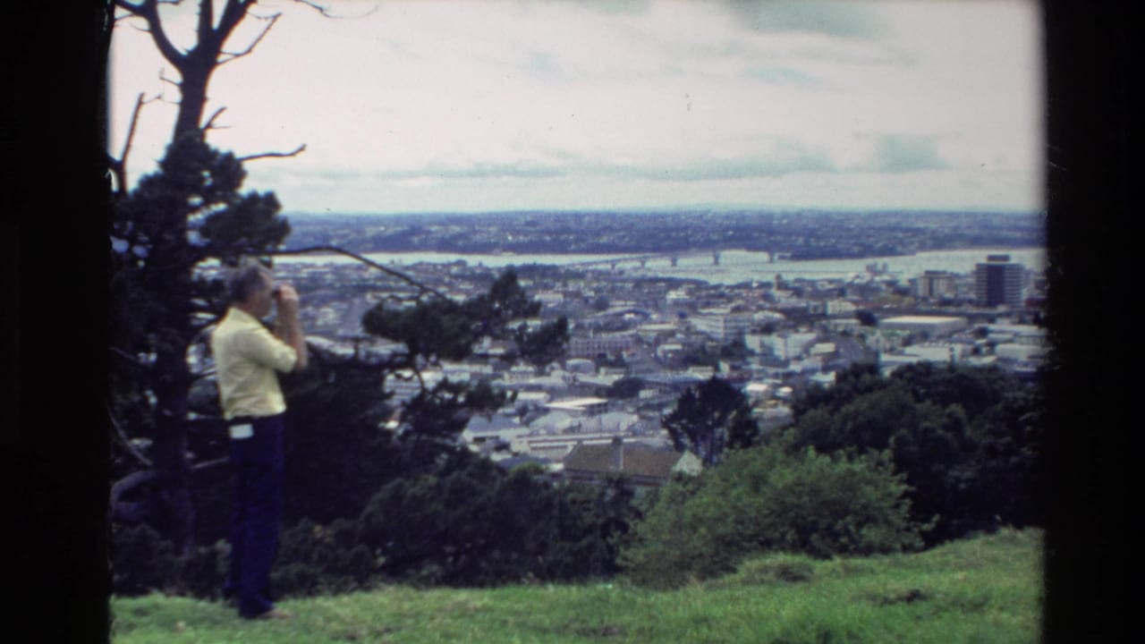 A man remains near the hilltop edge, looking through binoculars toward a New Zealand coastal town and harbor in 1981 footage.