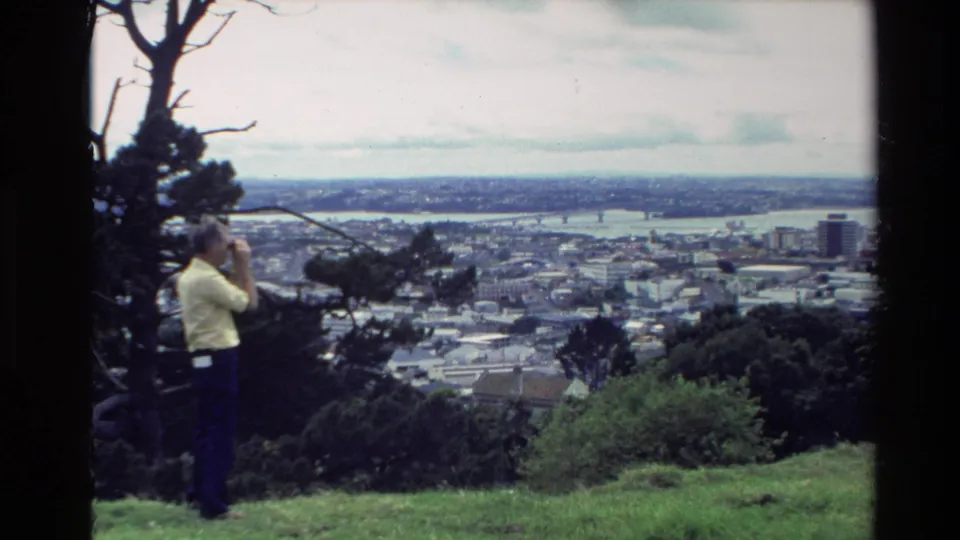 A man remains near the hilltop edge, looking through binoculars toward a New Zealand coastal town and harbor in 1981 footage.