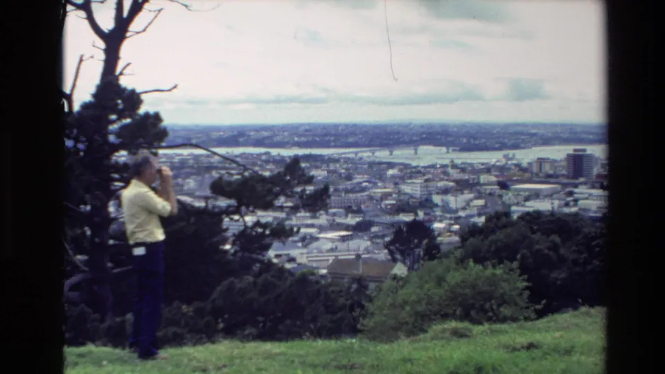 The 8mm frame shows the man still viewing the coastal town through binoculars, partly framed by trees and foreground grass.