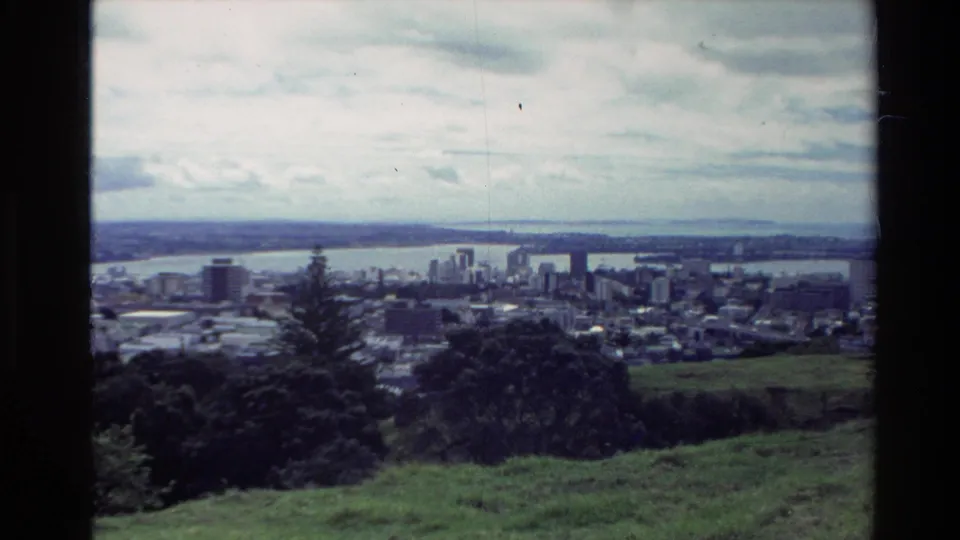1981 New Zealand 8mm frame looking from grassy hills toward a coastal city, harbor water, distant buildings, and a partly cloudy sky.