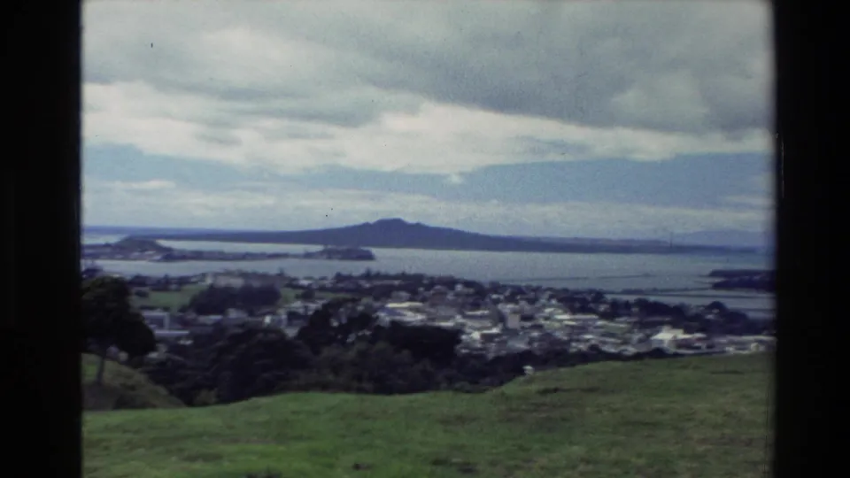 1981 New Zealand 8mm frame with rolling grass, dark foreground trees, coastal neighborhoods, harbor water, and distant land under clouds.