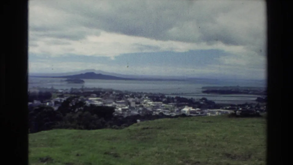 1981 New Zealand 8mm frame showing a broad harbor view from a green hillside, with urban buildings, shoreline, and distant landforms.