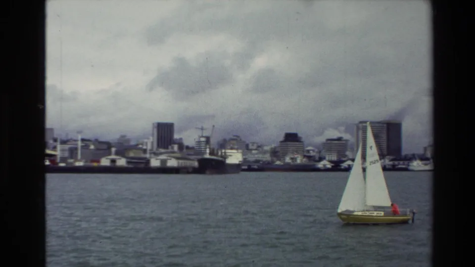A yellow-hulled sailboat with white sails crosses a New Zealand urban bay in 1981, with mid-rise buildings and harbor structures behind it.