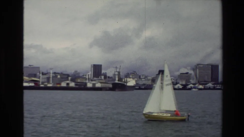 The sailboat continues across the New Zealand bay, framed against distant waterfront buildings, cranes, and a gray clouded sky.
