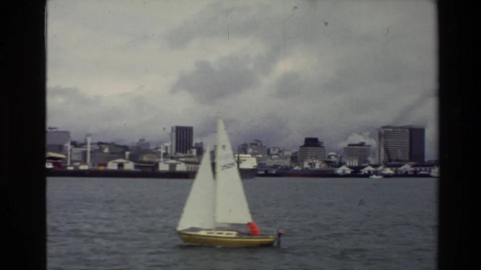 A closer 1981 home-movie view shows the yellow-hulled sailboat under white sails with an urban New Zealand waterfront behind it.