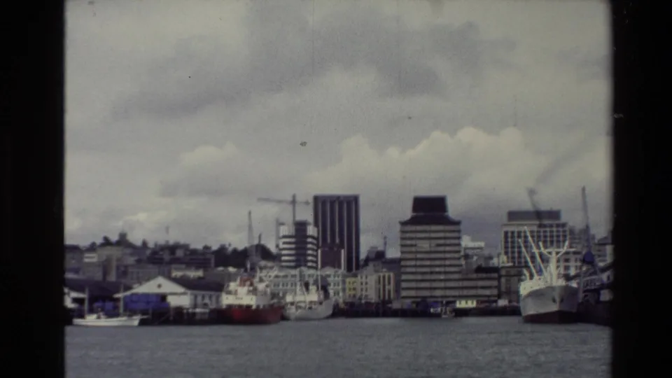 1981 New Zealand harbor view with moored vessels, dock cranes, waterfront buildings, and muted 8mm color from across the water.