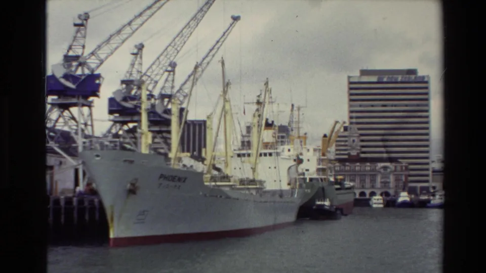 A large cargo vessel marked PHOENIX is docked at a New Zealand port in 1981, with cranes, tugboats, and waterfront buildings behind it.