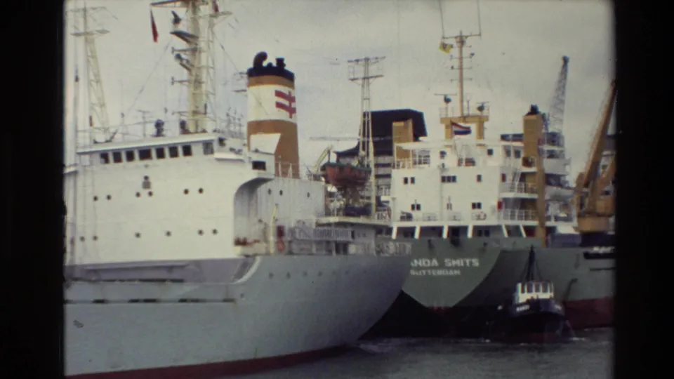 Close 8mm view of cargo ships at a New Zealand harbor, including a stern marked MANDA SMITS with cranes and a small tugboat nearby.
