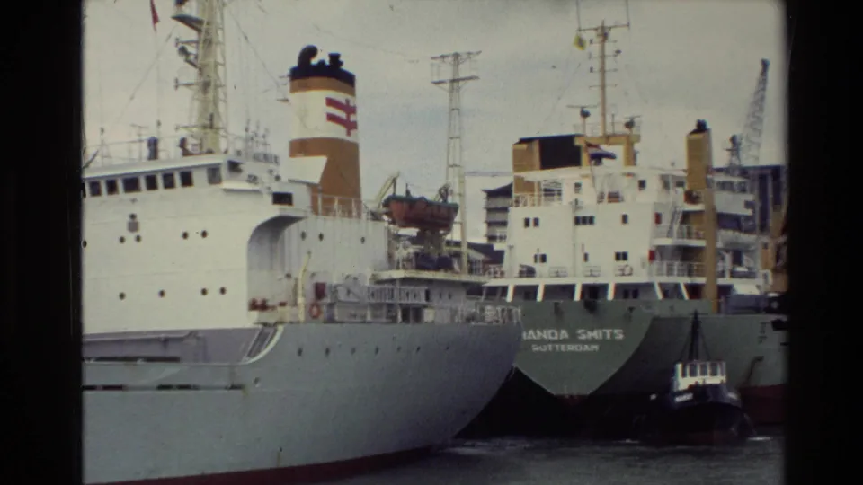 The stern of MANDA SMITS appears in a 1981 New Zealand harbor scene, with another large ship, funnel markings, cranes, and a small tugboat visible.