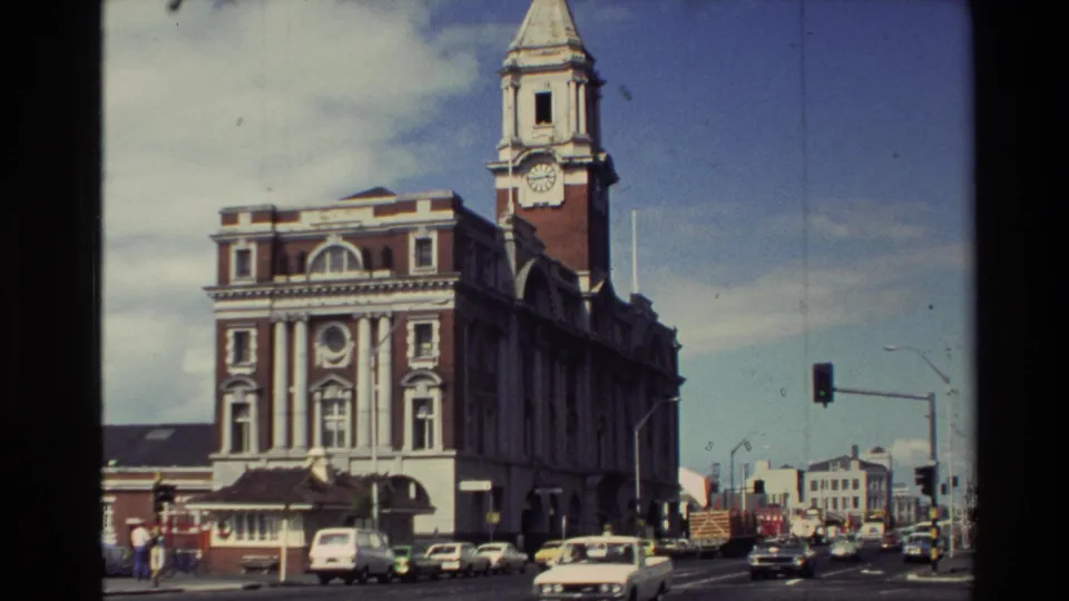 1981 New Zealand street view with a prominent clock tower building, moving cars, traffic lights, and pedestrians near the curb.