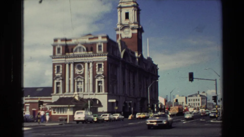 1981 New Zealand city street scene with a clock tower building, multiple cars, pedestrians on the sidewalk, and traffic lights.