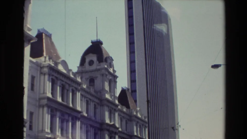 Handheld 8mm frame of the older building and adjacent tower, with the camera angled upward from the city street.