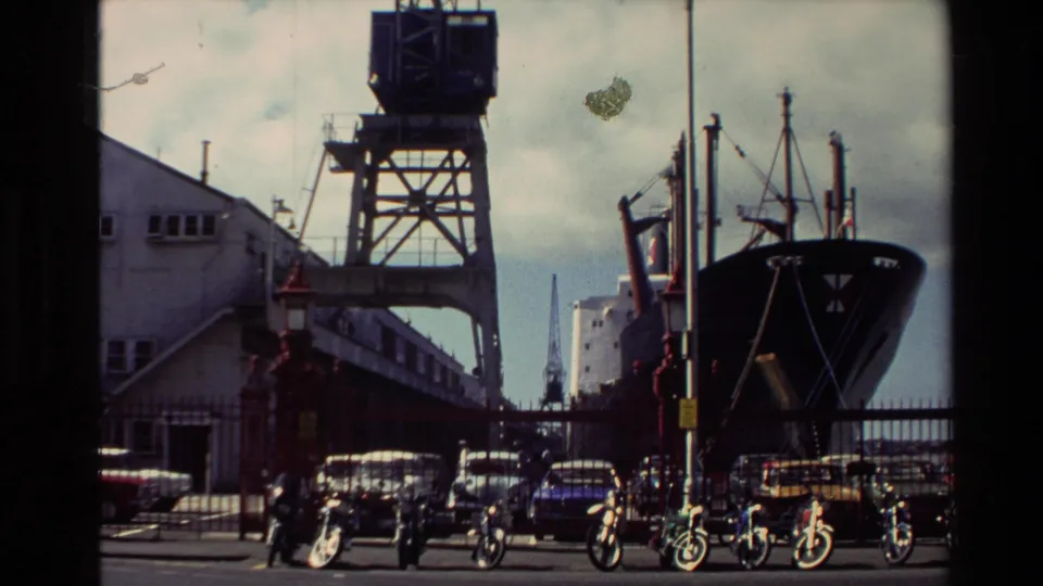 1981 New Zealand waterfront view with a docked ship, port crane, parked motorcycles, cars, and industrial buildings.