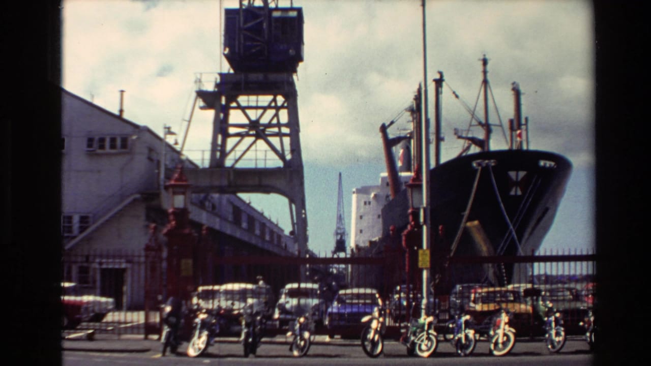 1981 New Zealand harbor view with a cargo ship beside cranes, parked cars, motorcycles, and a white dockside building.