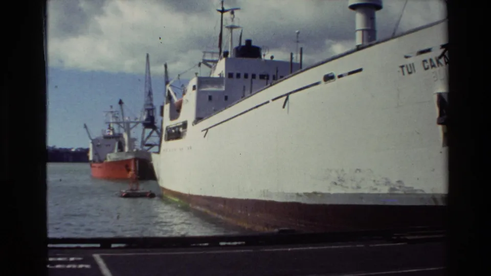 White vessel marked with visible lettering docked in a New Zealand harbor, with another ship and cranes behind it.