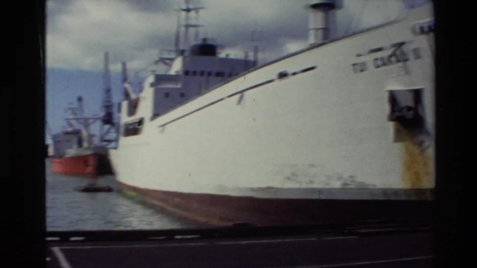 1981 New Zealand 8mm frame showing the side of a large white cargo ship at harbor, with rust staining and a red tugboat behind it.