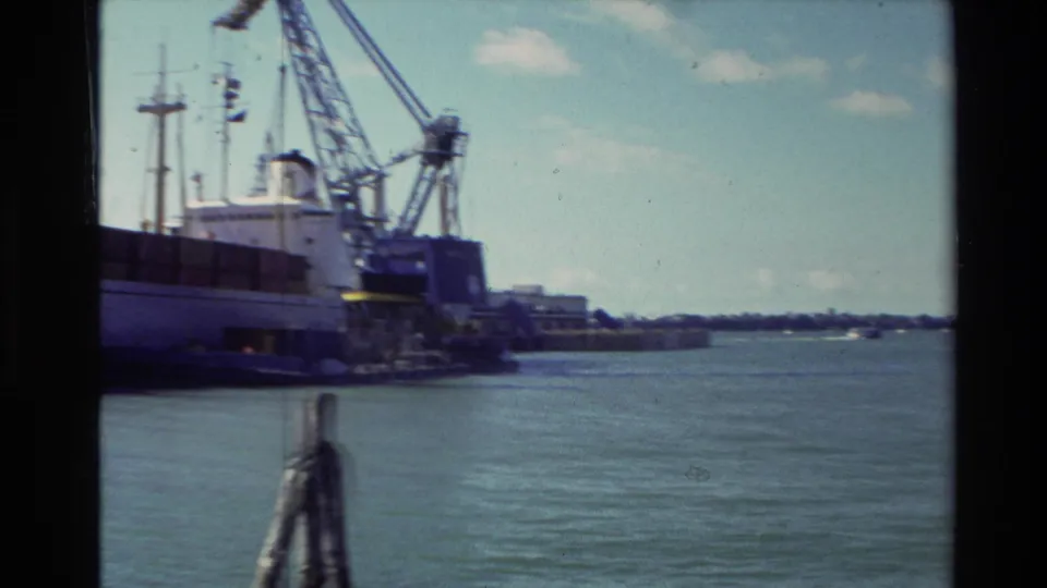 1981 New Zealand 8mm frame showing a cargo ship and cranes at harbor, with open water and distant shoreline beyond.