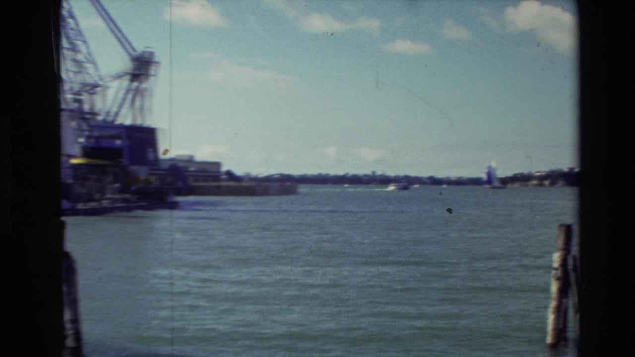 1981 New Zealand 8mm frame showing water and distant shoreline through dark window bars, with a crane-like structure at the far left.