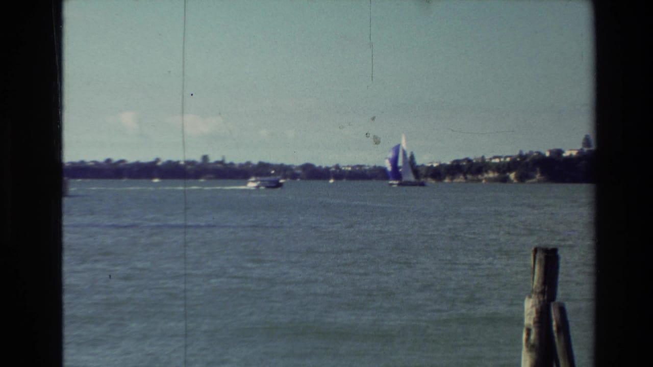1981 New Zealand 8mm frame of a lagoon or harbor with small boats and a blue-and-white sailboat near the right edge.