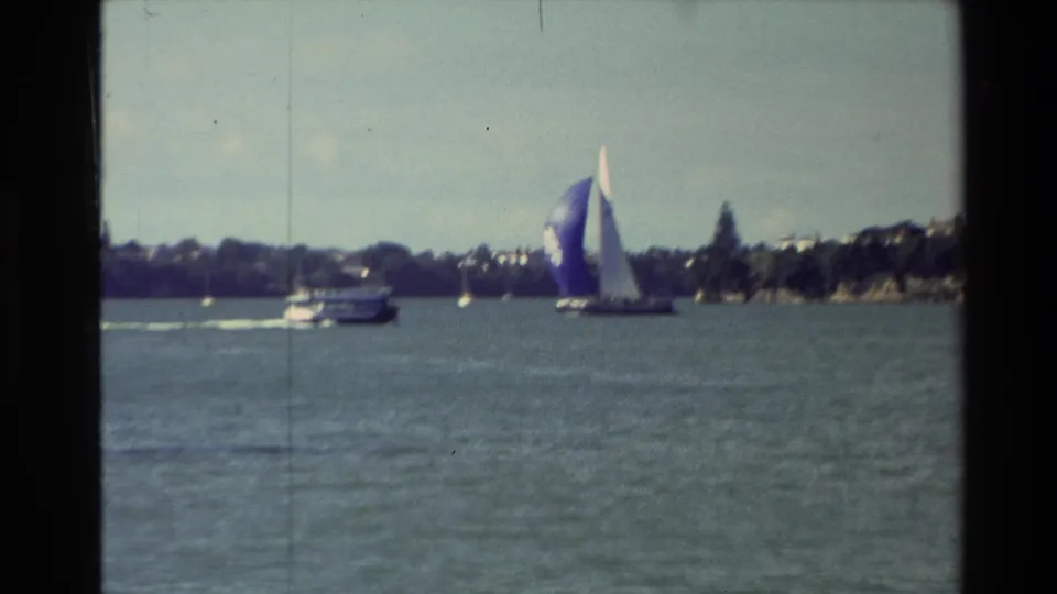 1981 New Zealand 8mm frame with a blue-and-white sailboat crossing near a motor boat, backed by a tree-lined shoreline.