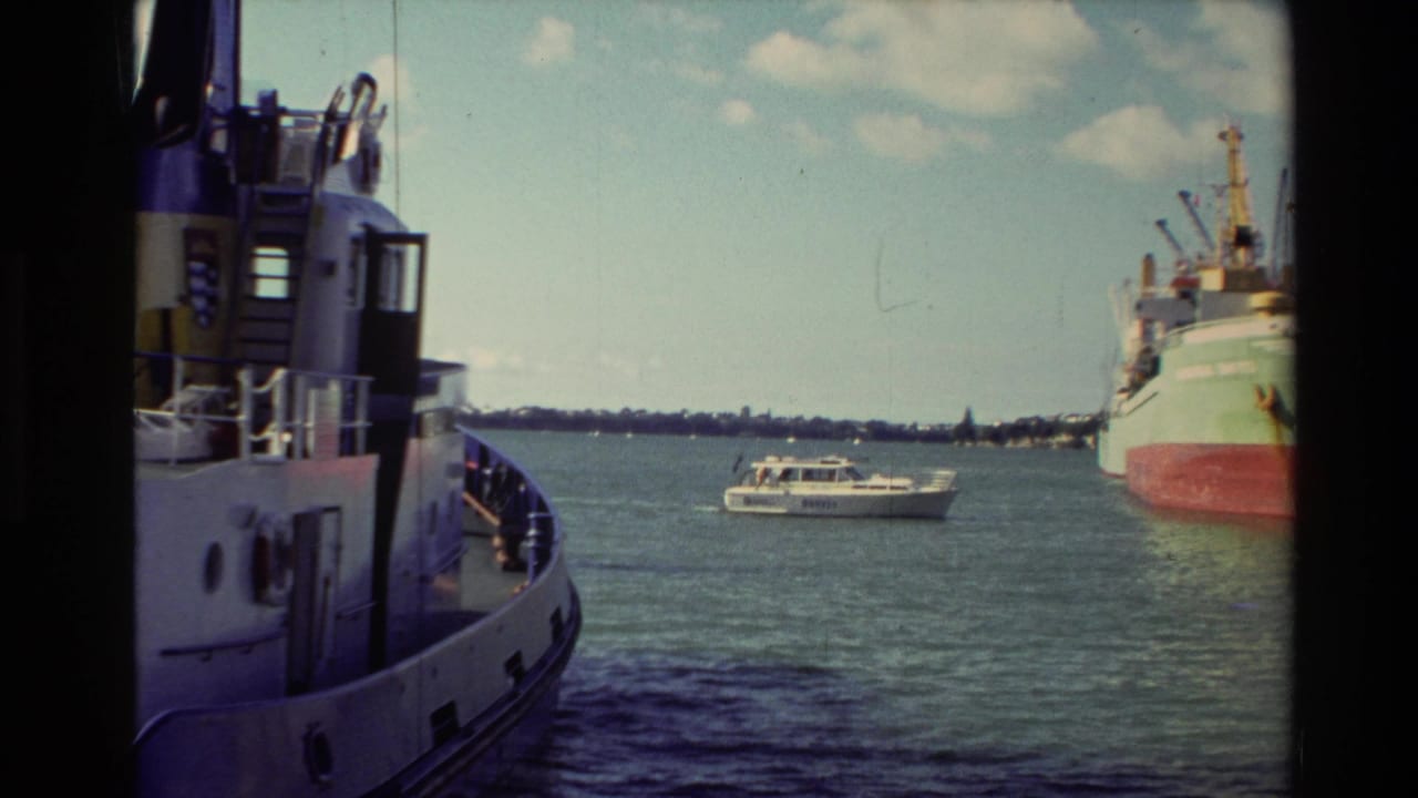 1981 New Zealand harbor footage showing a docked cargo ship, a white motorboat, distant shoreline, and the side of a nearby vessel.