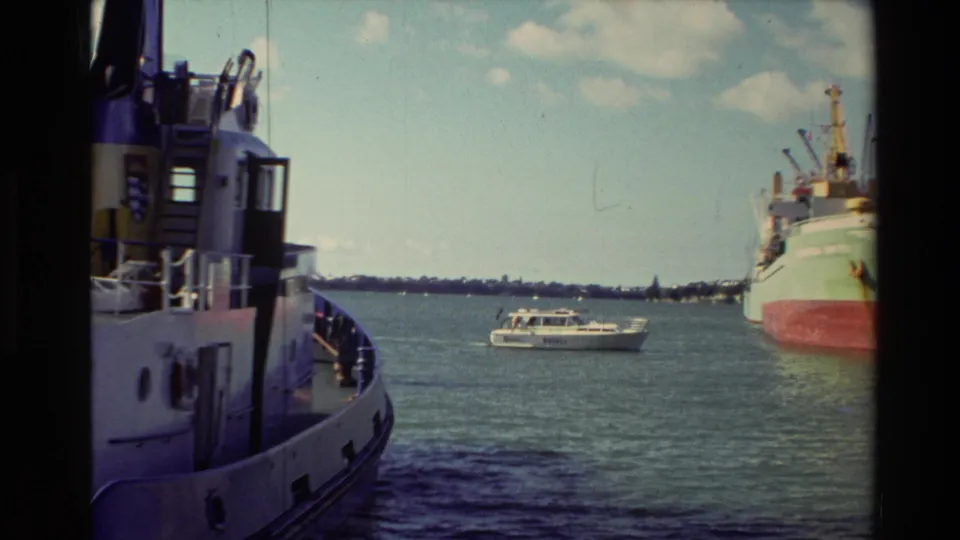 1981 New Zealand harbor footage showing a docked cargo ship, a white motorboat, distant shoreline, and the side of a nearby vessel.