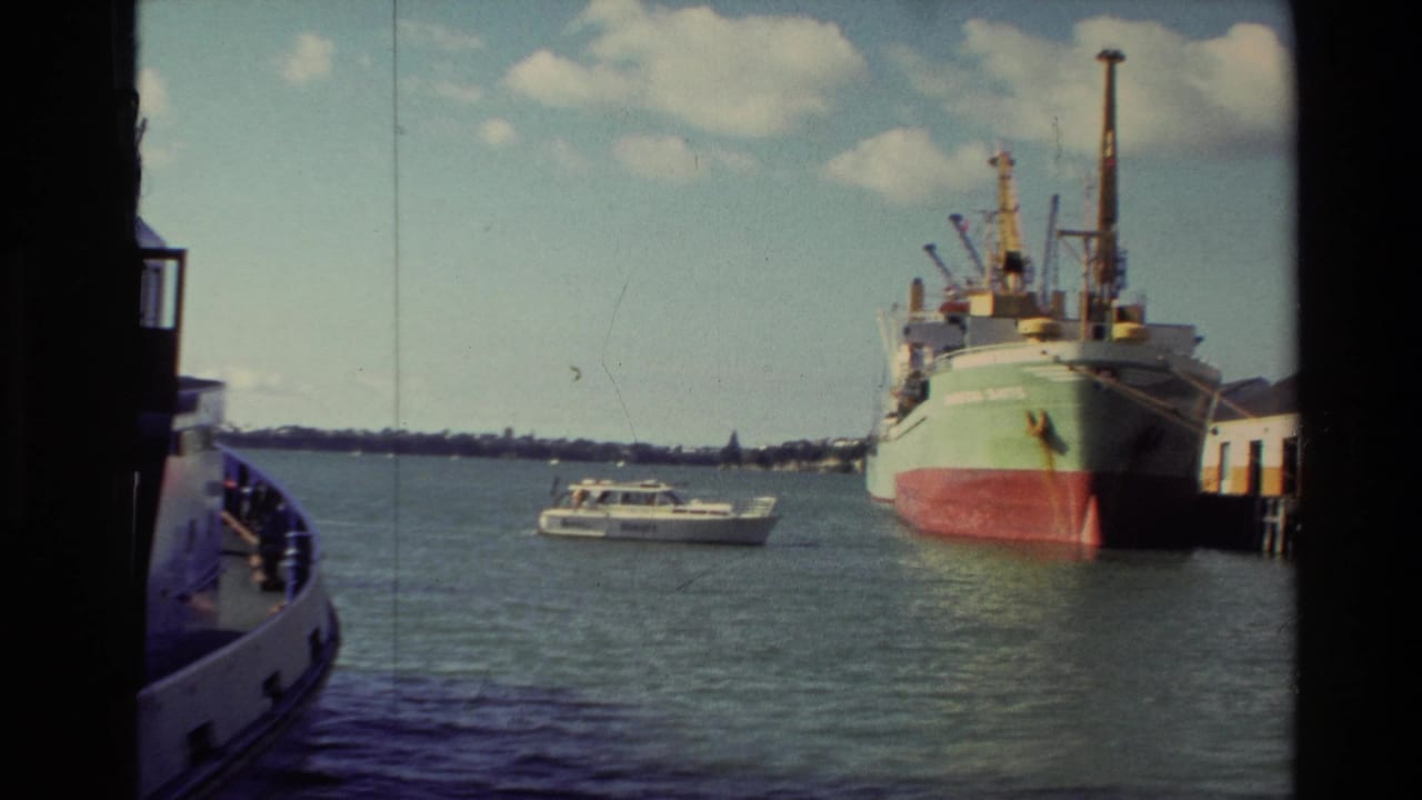 1981 New Zealand harbor scene with a large cargo ship at a pier, a white motorboat on the water, and dock structures at right.