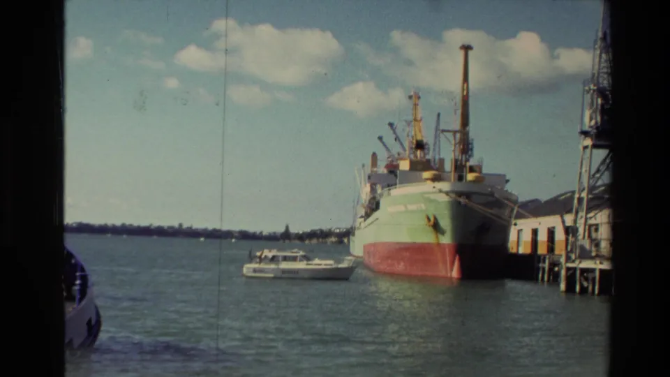 1981 New Zealand harbor frame with a moored cargo ship, dockside structures, small motorboat, calm water, and distant low shoreline.