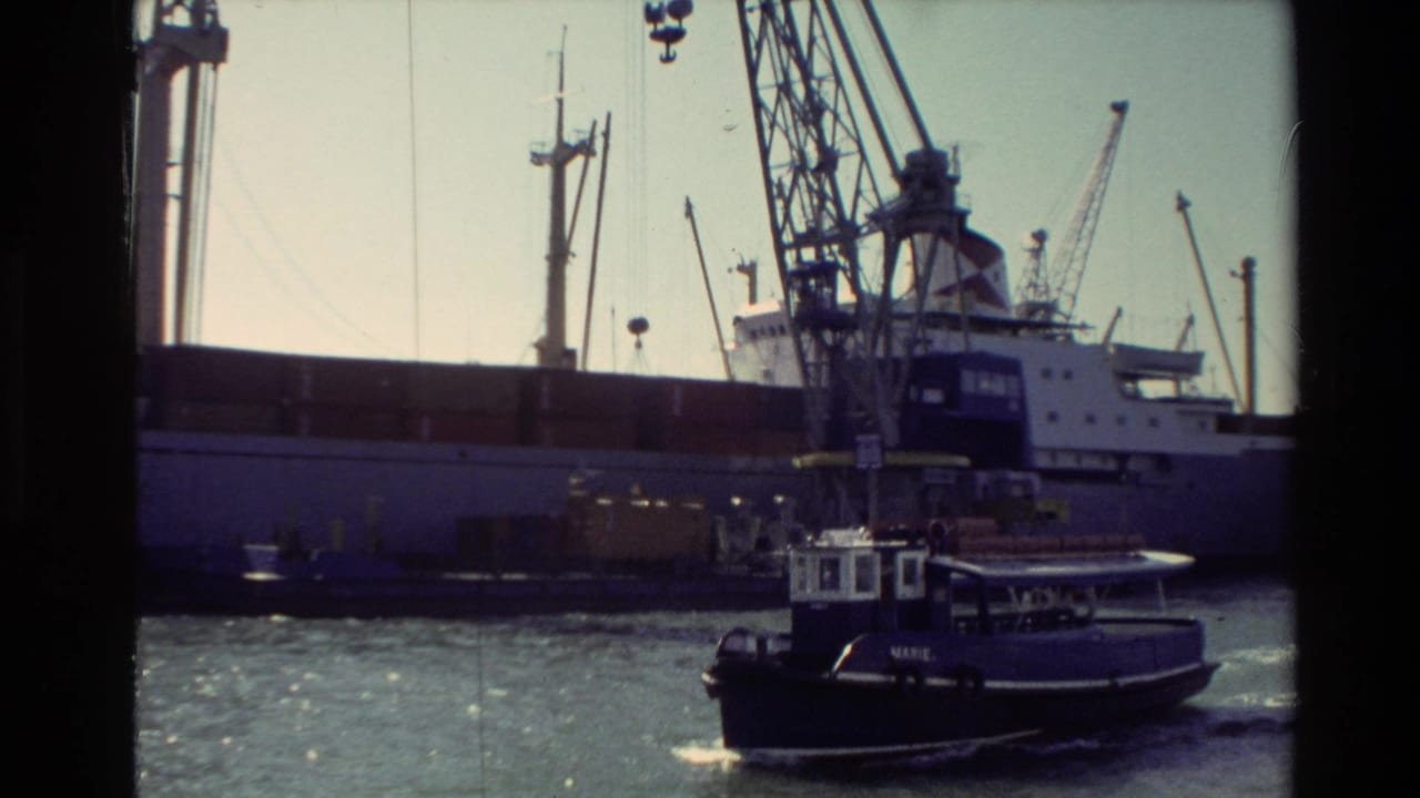 1981 New Zealand harbor view with a tugboat passing a large barge beneath tall industrial cranes.