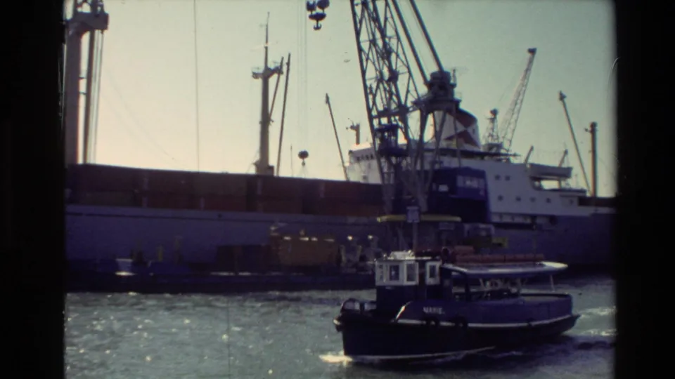 1981 New Zealand harbor view with a tugboat passing a large barge beneath tall industrial cranes.