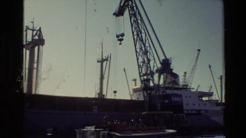 1981 New Zealand industrial waterfront view with tall cranes, a docked barge, and sunlit harbor water.