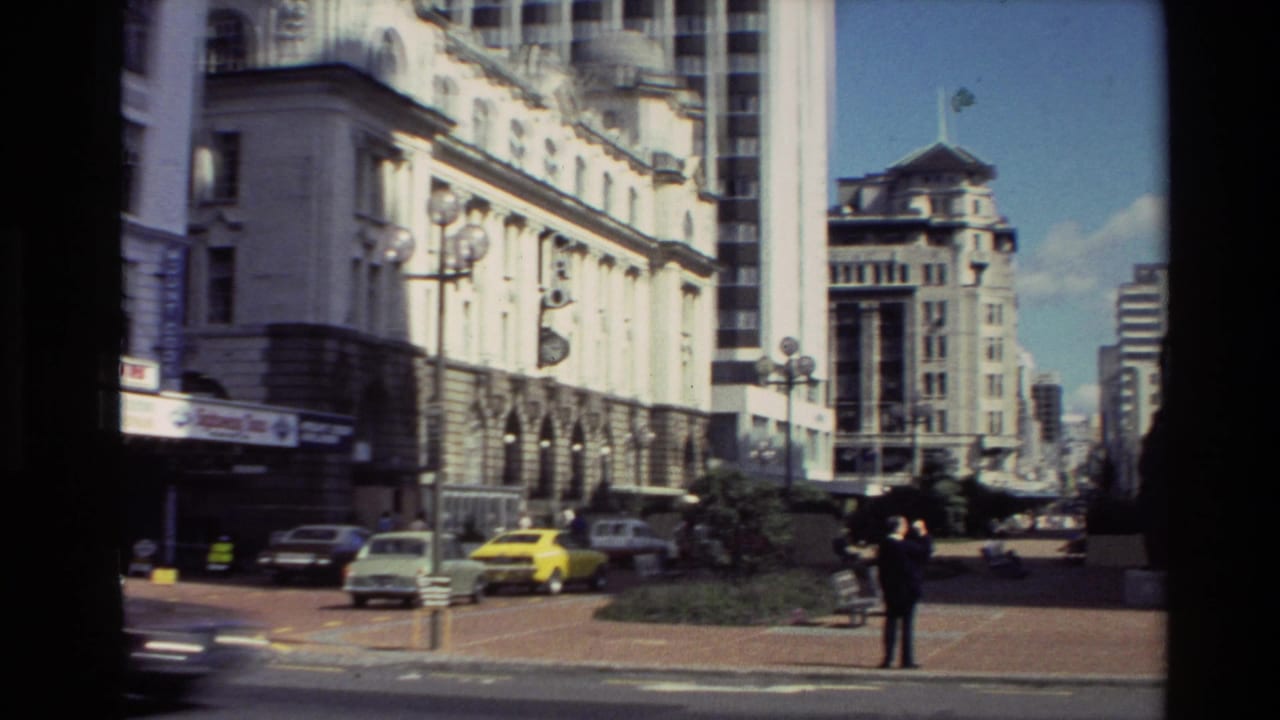 City street with parked cars, a yellow sedan, tall buildings, and a man standing on the sidewalk.