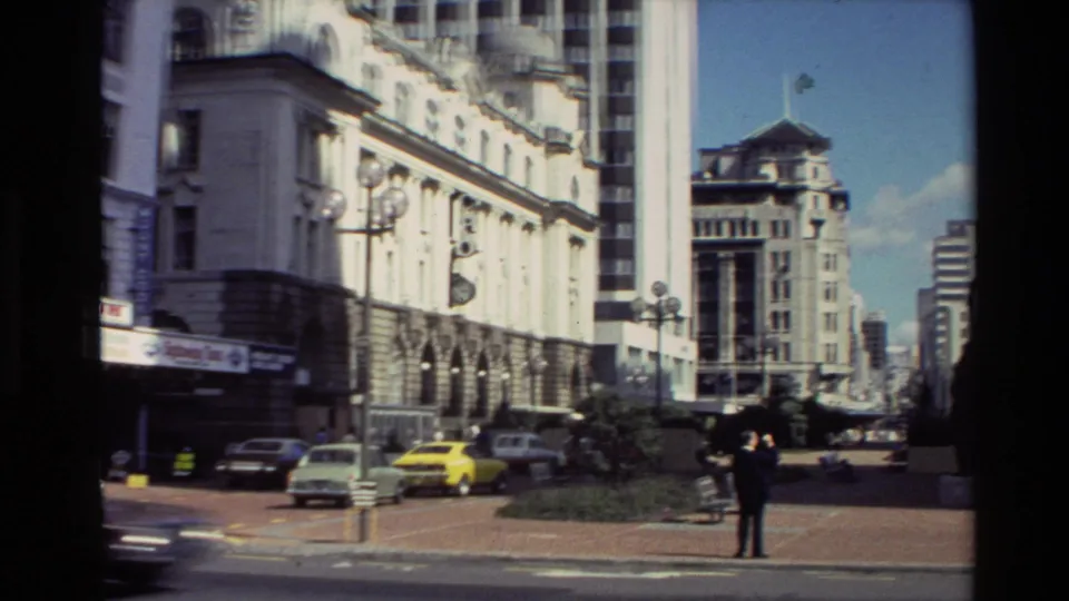 1981 New Zealand street view with parked classic cars, a bright yellow sedan, a suited man, historic facades, and taller city buildings.