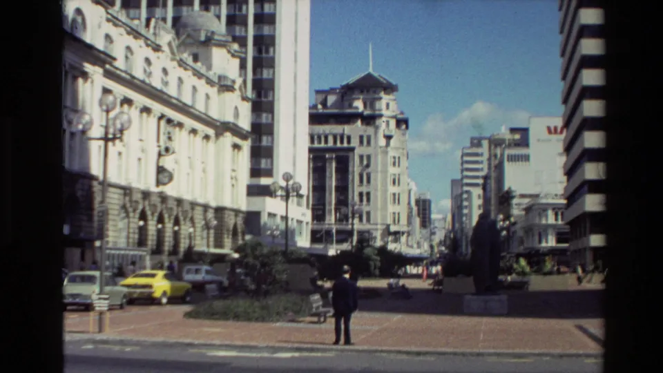 1981 New Zealand 8mm street frame showing a suited figure on the pavement, parked cars, ornate lamps, and downtown architecture.