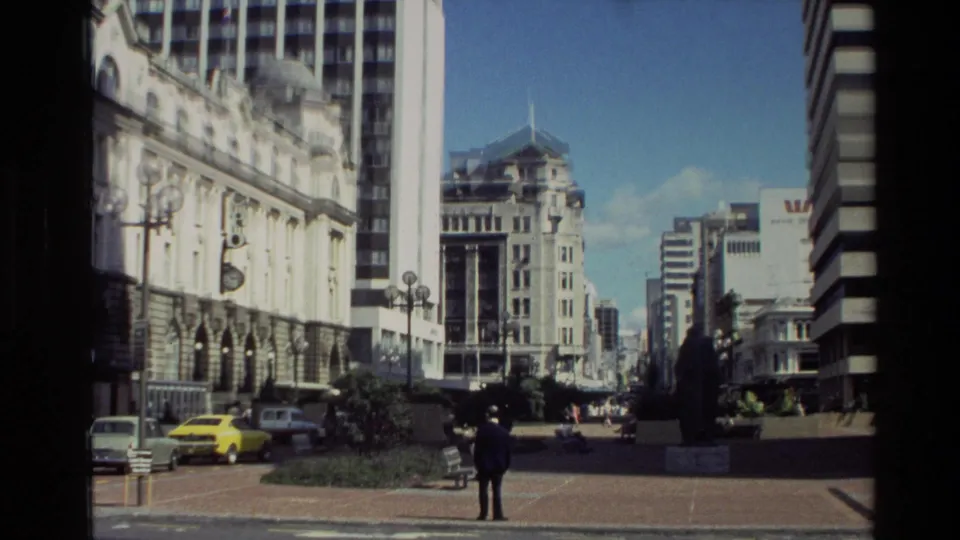 1981 New Zealand city view with a suited man in the foreground, a yellow car at the curb, historic masonry, and modern towers beyond.