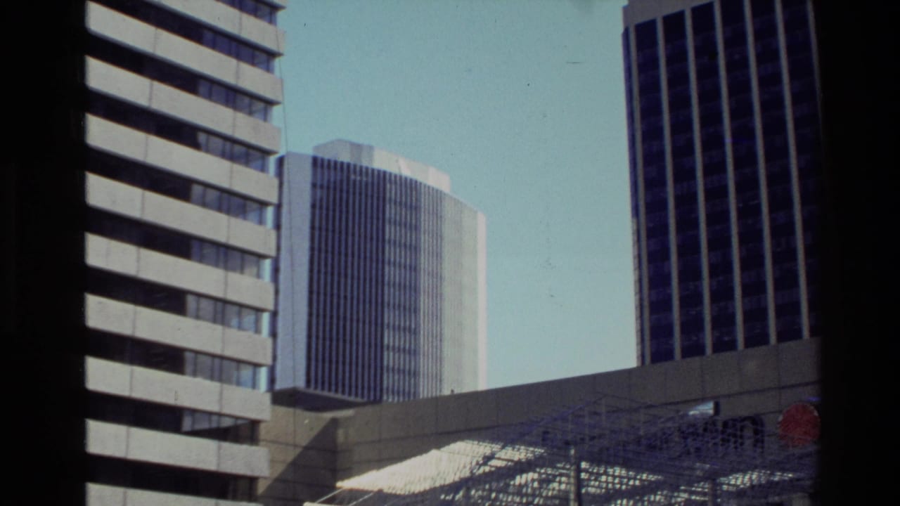 1981 New Zealand home-movie frame showing high-rise buildings over a modern metal plaza sculpture or canopy.