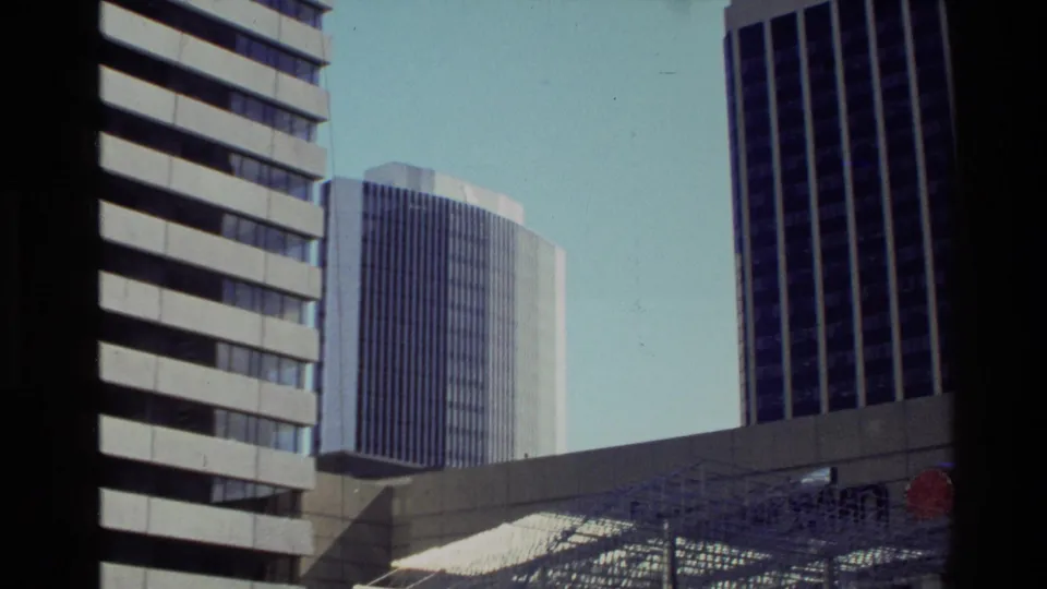 1981 New Zealand home-movie frame showing high-rise buildings over a modern metal plaza sculpture or canopy.