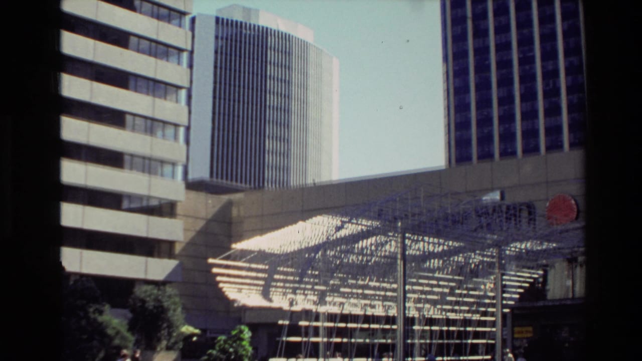 1981 New Zealand city plaza view with office towers, a geometric metal installation, and an unreadable red sign.
